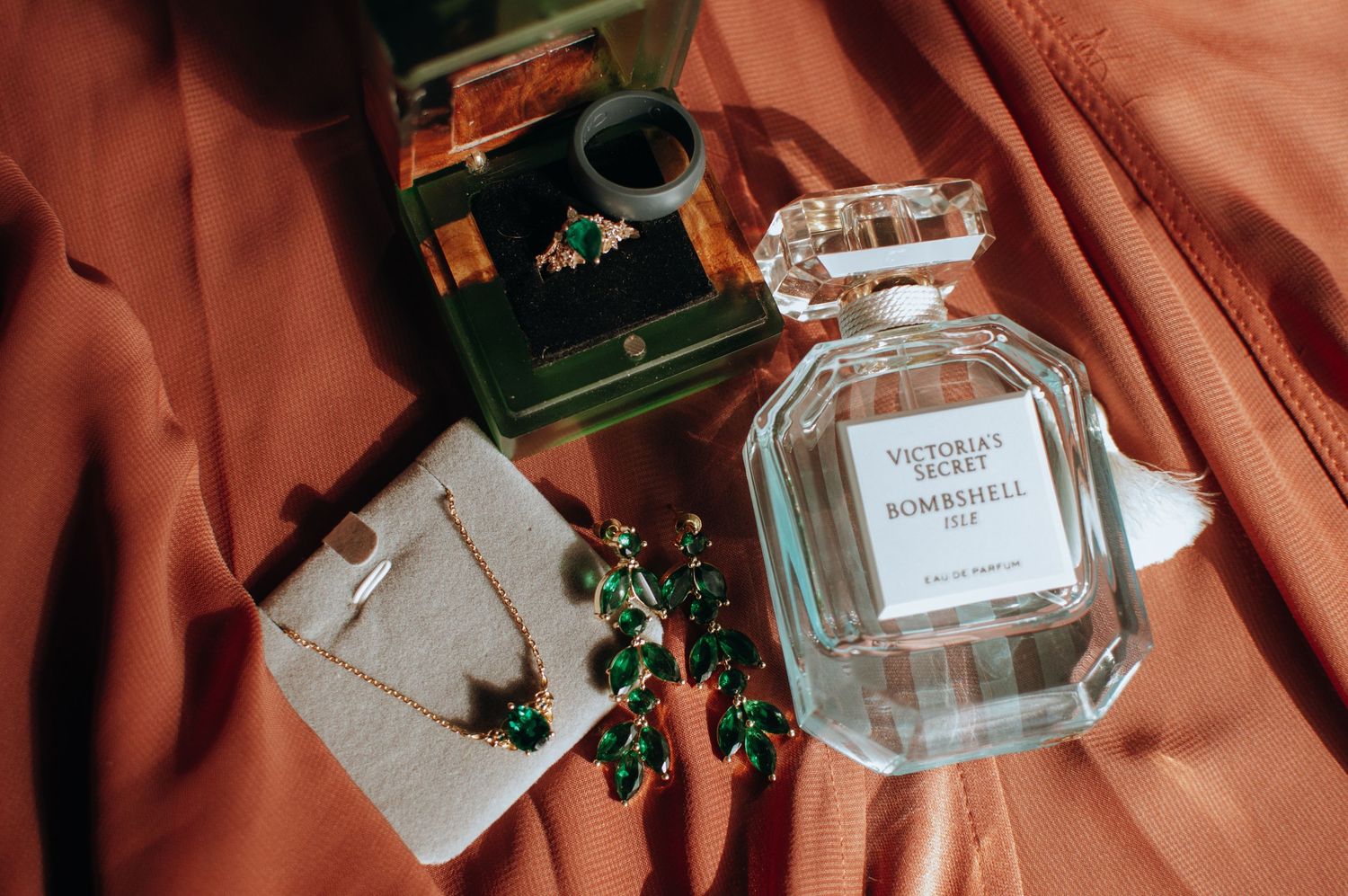 Glass perfume bottle and delicate jewelry displayed on coral-colored fabric with green accents.