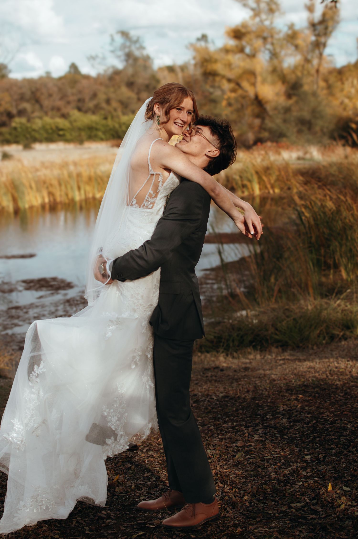 A romantic wedding moment captured by a lake with autumn foliage in the background.