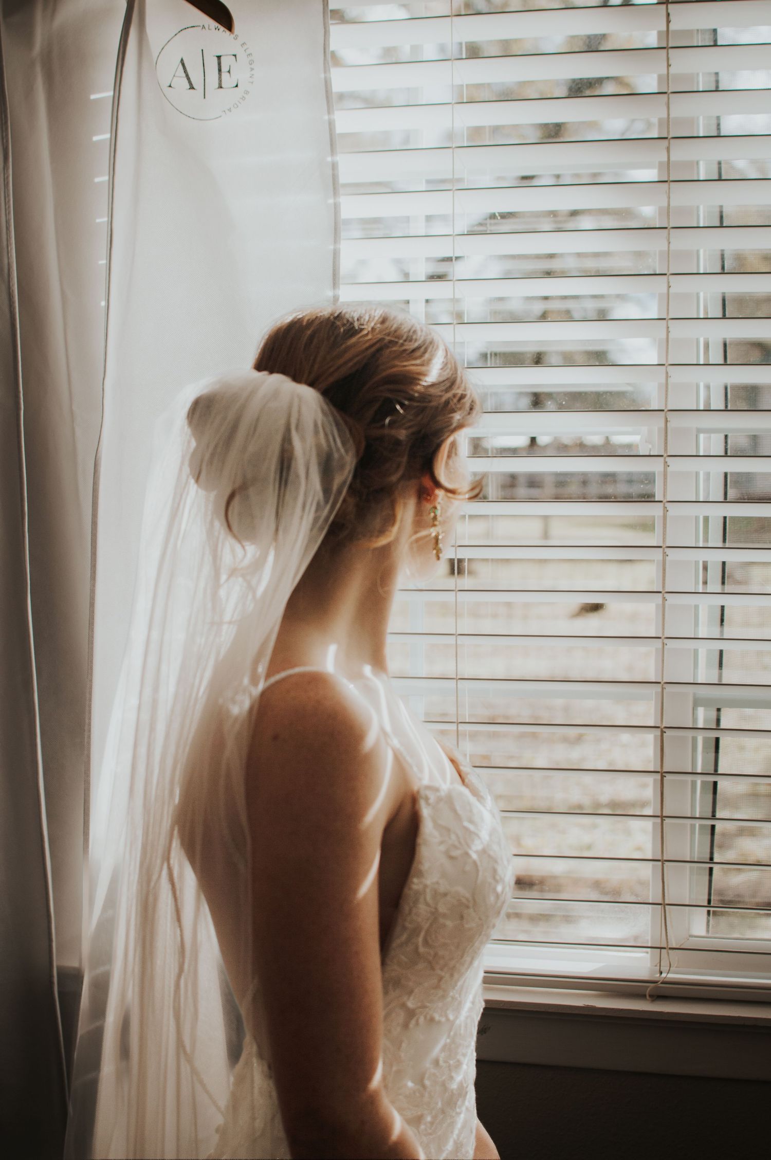 Silhouette of bride in lace dress gazing through window blinds.