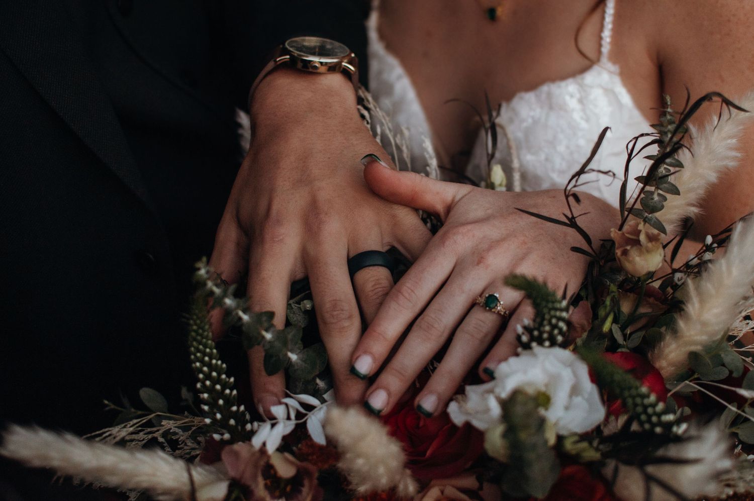 Close-up detail of wedding rings with dried flowers and feathers in a moody dark setting.