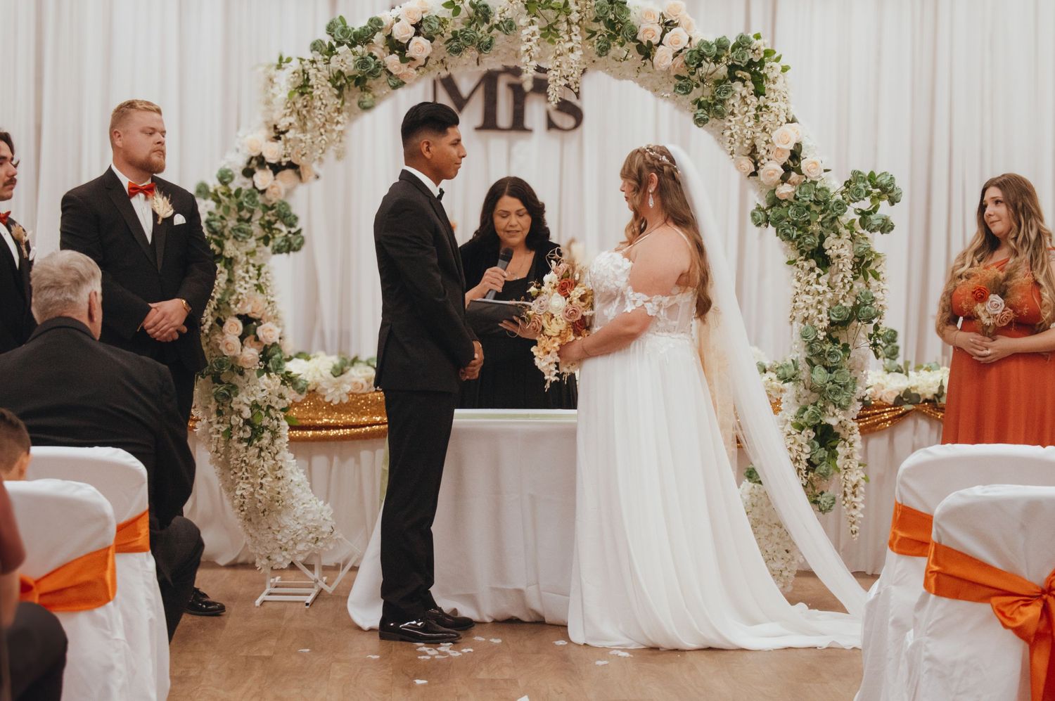 A wedding ceremony takes place under an elegant floral arch with 'Mrs' lettering and orange chair accents.
