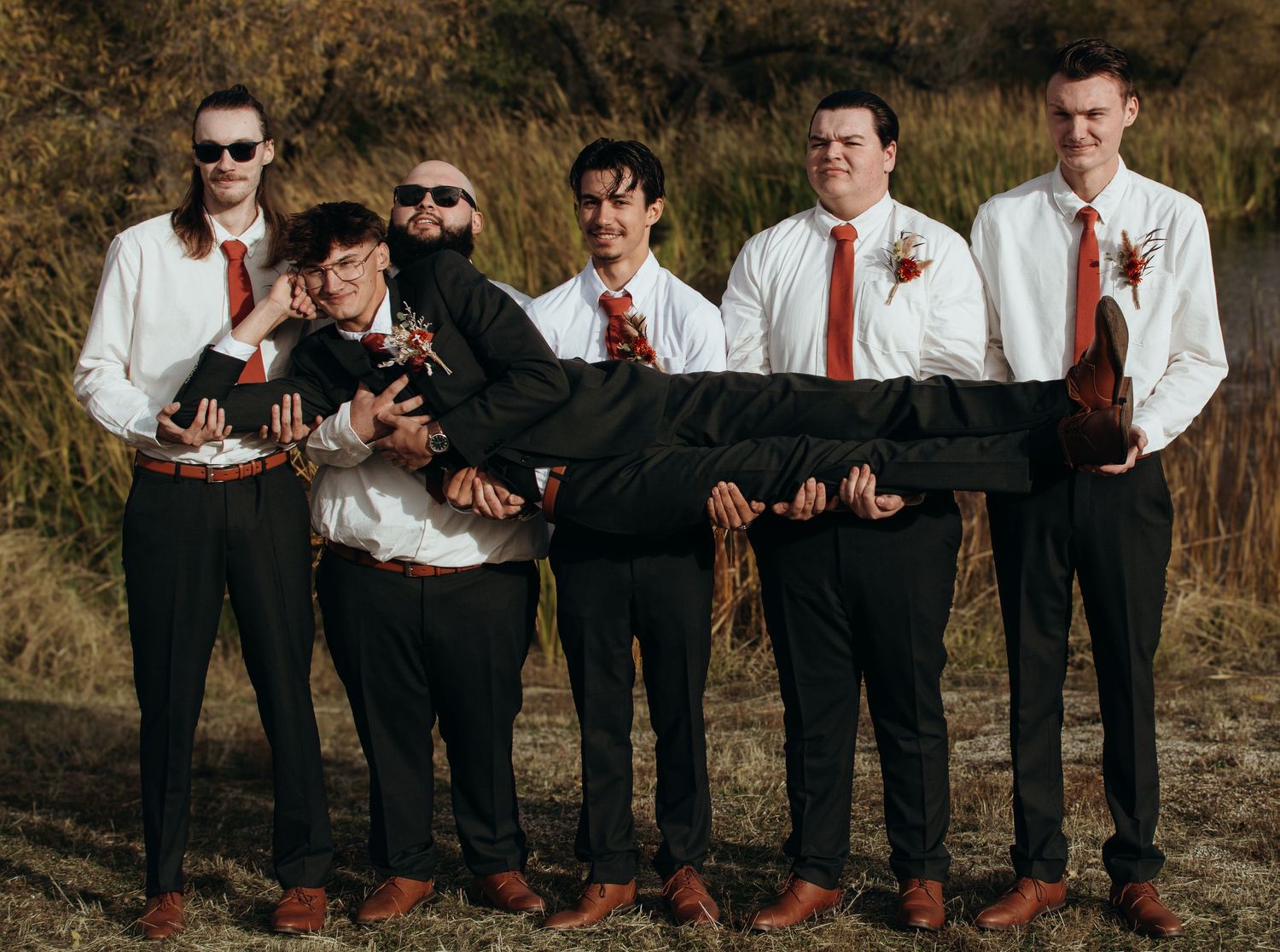 Groomsmen in matching attire pose playfully while lying in a row on fallen autumn leaves.