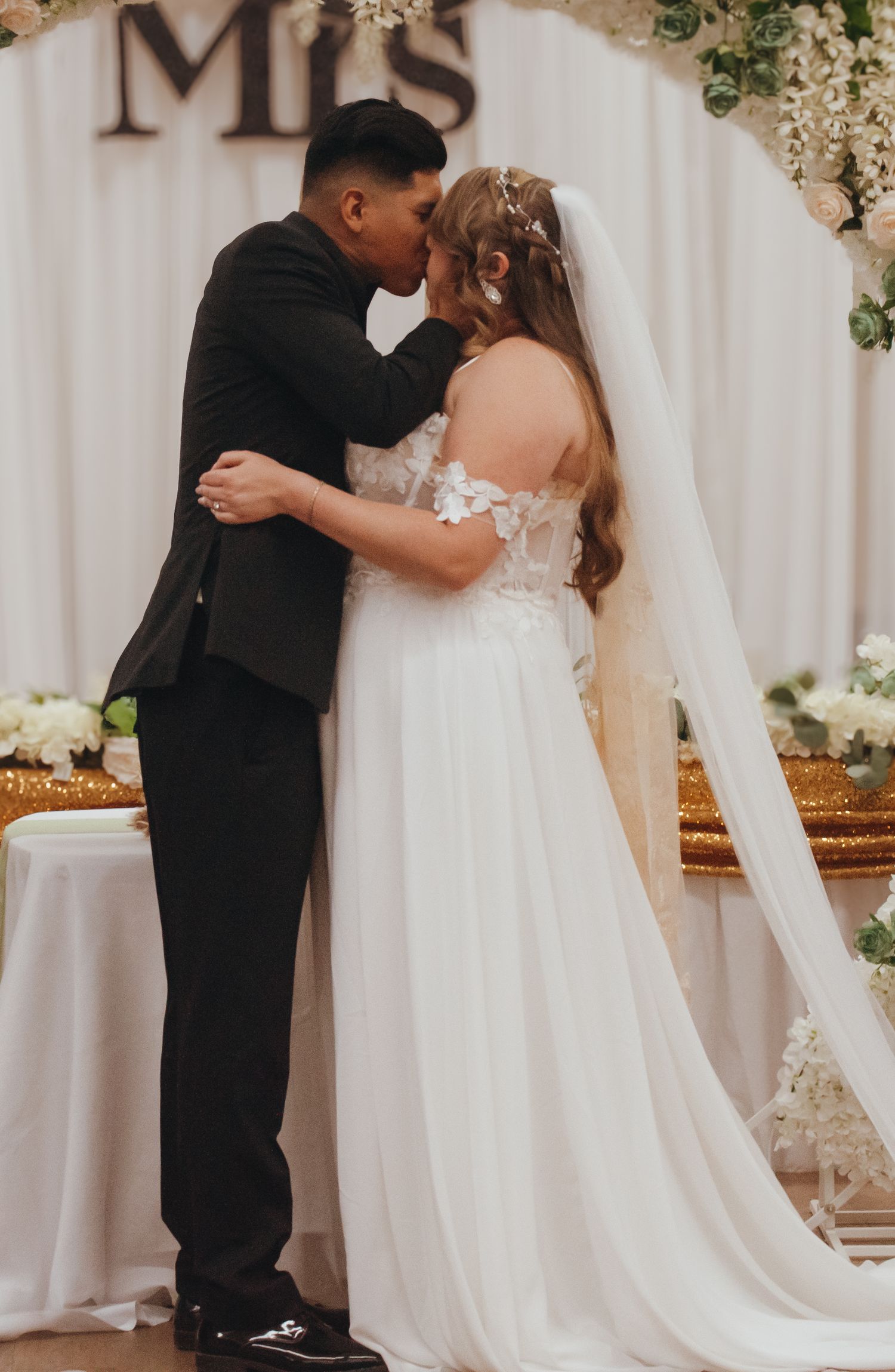 A romantic wedding moment captured under a floral arch with 'Mrs' signage in the background.