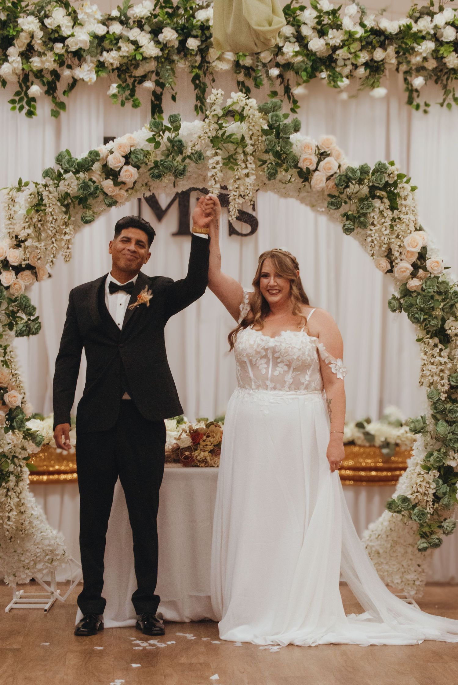 Newlyweds celebrate under a floral arch decorated with white roses and greenery.