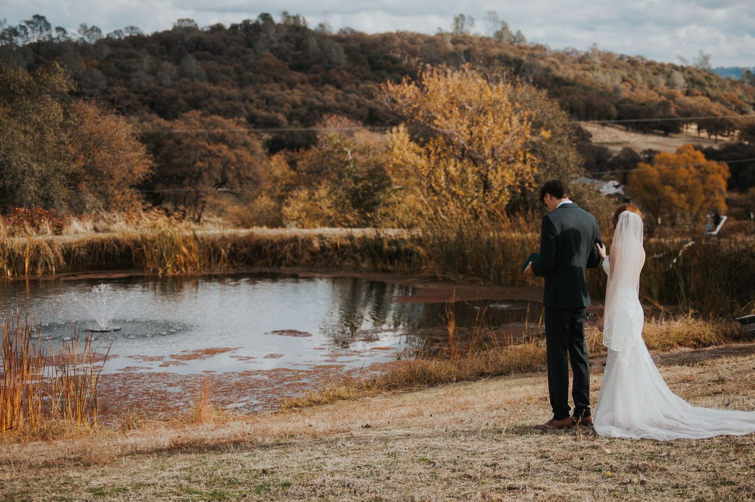 Wedding couple stands together against scenic autumn landscape with water and rolling hills.