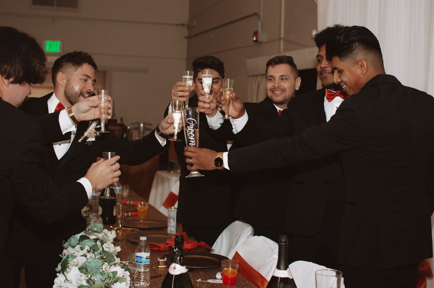 Group of men in formal wear toasting with bottles at a wedding reception table.