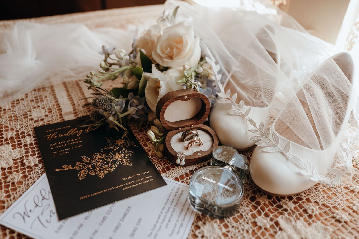 Wedding details flat lay with invitation, rings, and white roses on sequined fabric.