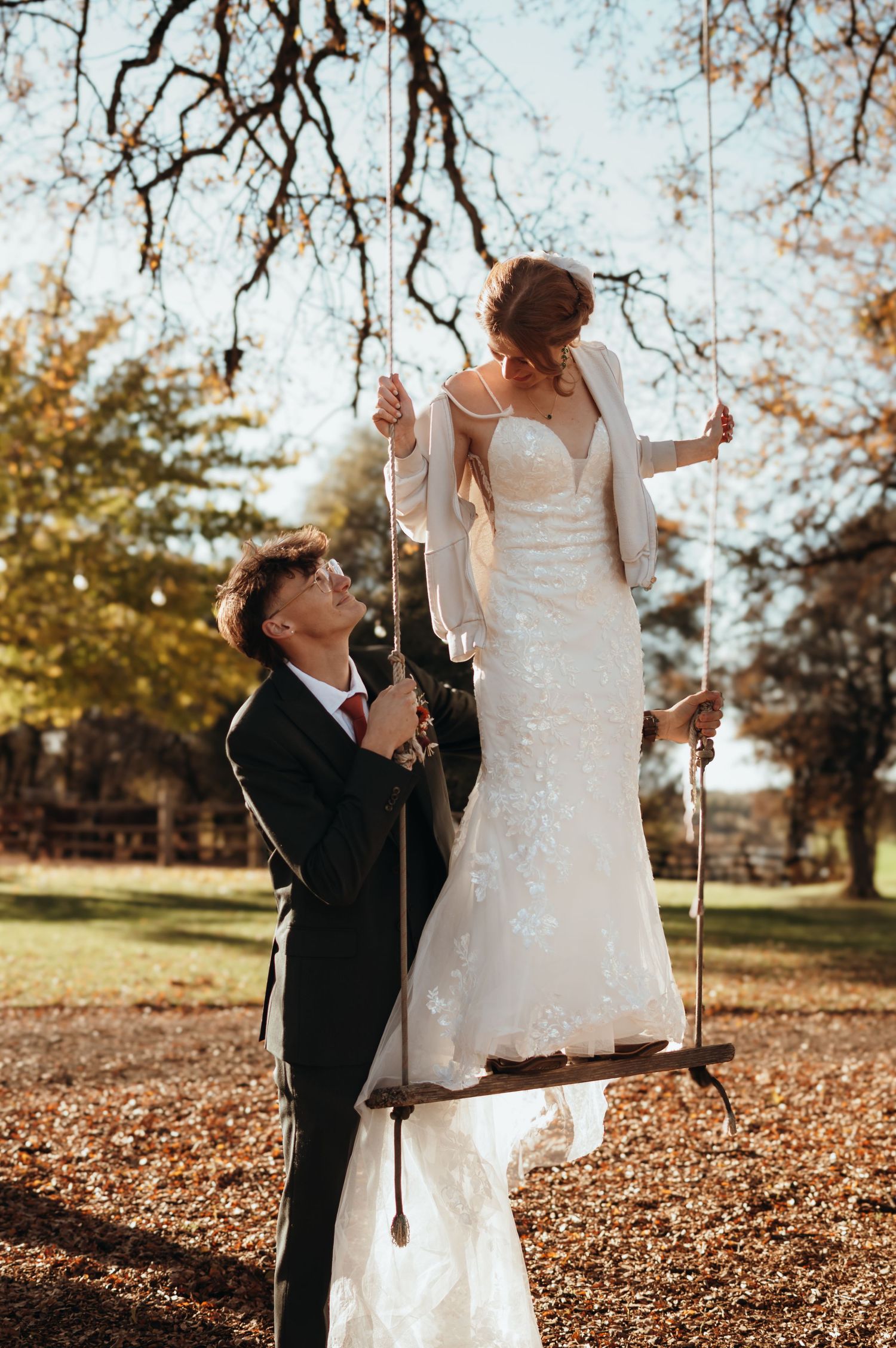 Playful autumn wedding scene with couple among fallen leaves under bare tree branches.