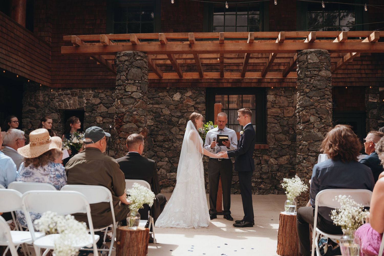 Outdoor wedding ceremony under wooden pergola with stone pillars and white chairs.