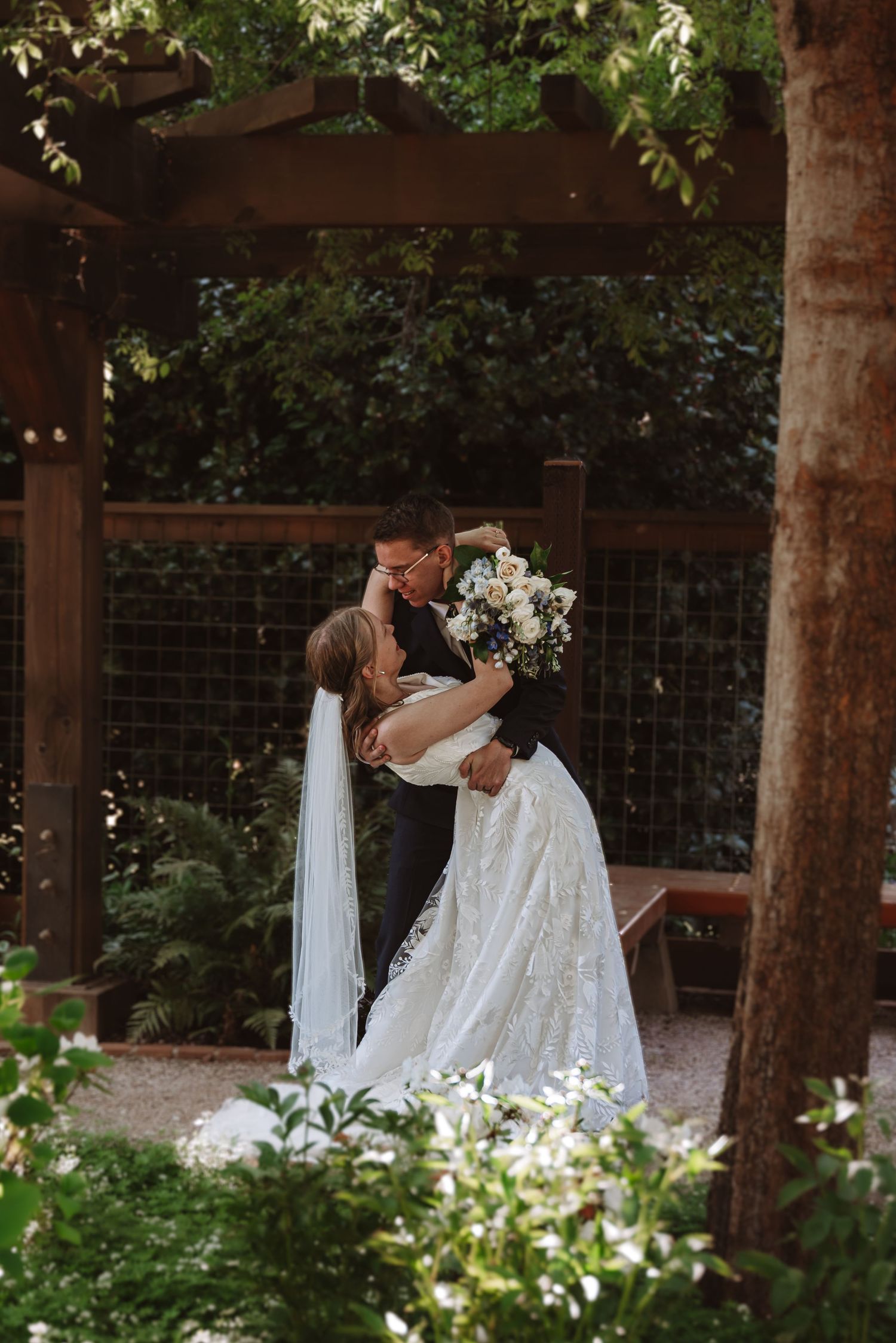 A couple shares their first dance under a wooden pergola decorated with white fabric and greenery.