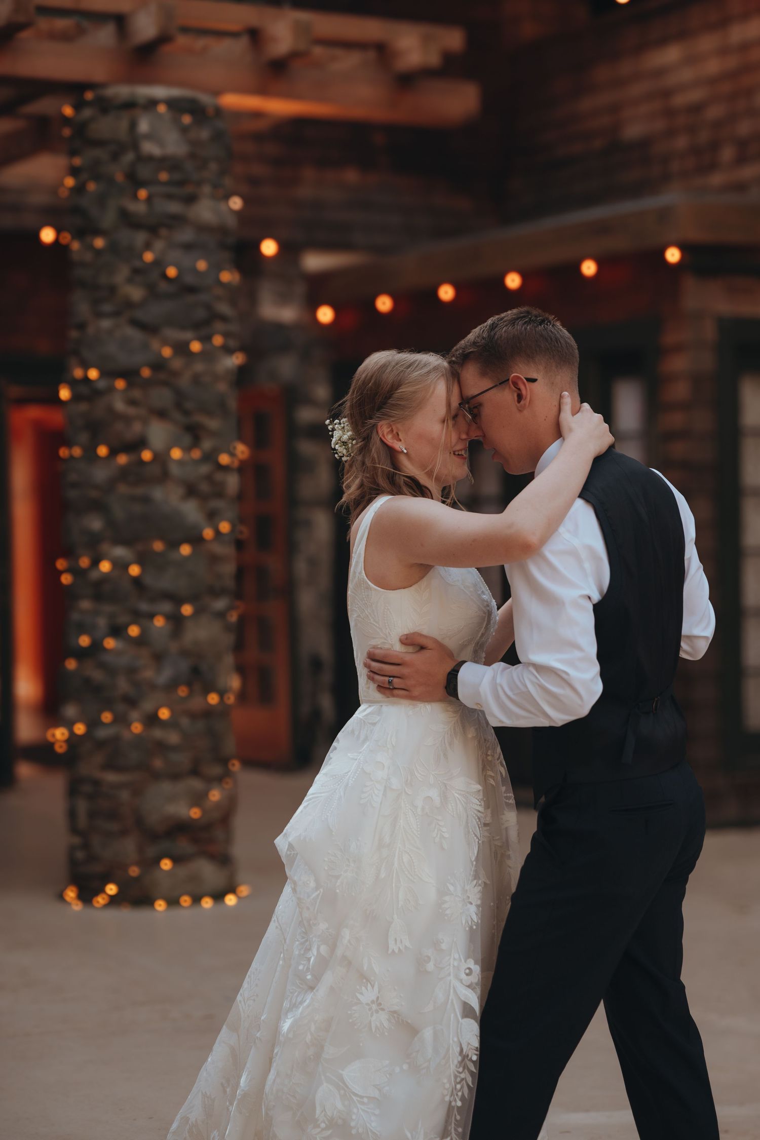 A couple dances intimately in a rustic venue decorated with string lights.