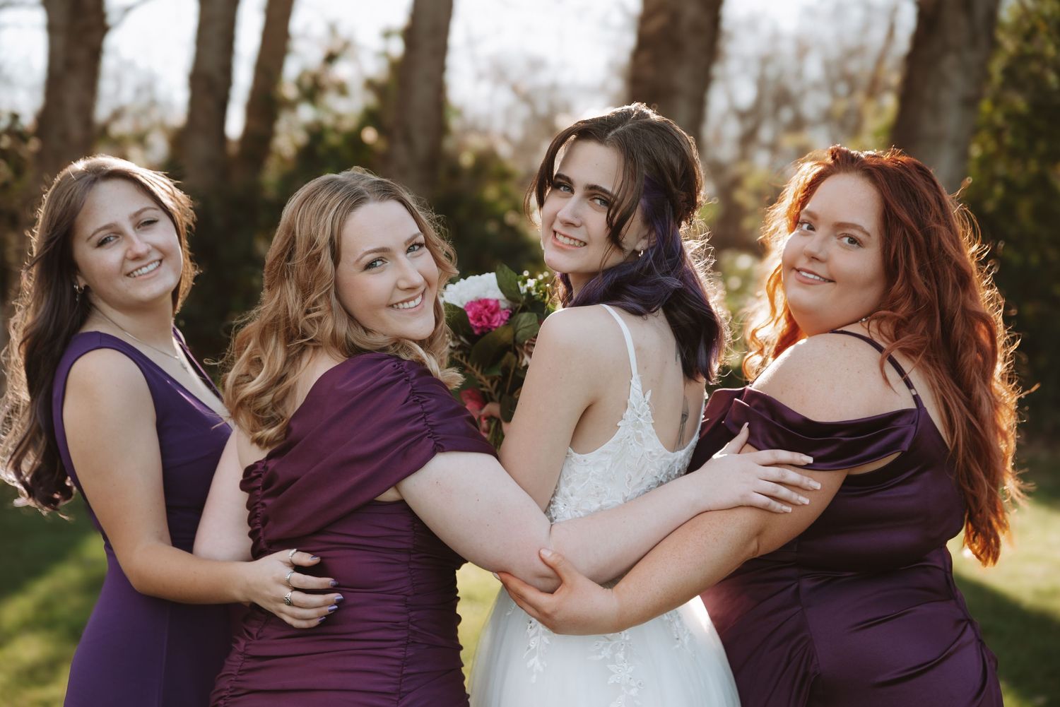 Bridesmaids in purple dresses share a joyful group hug with the bride in white.
