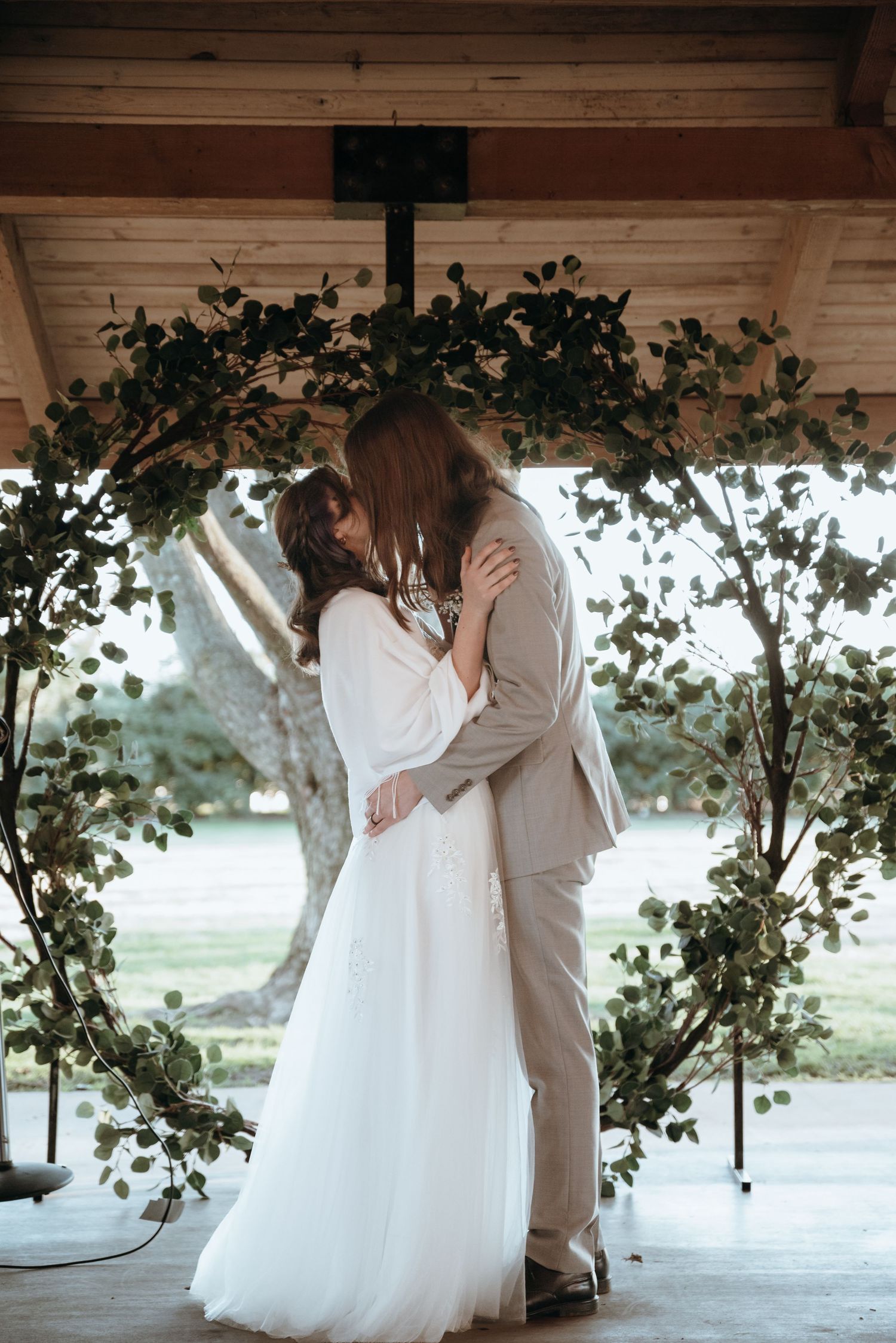 A couple shares a tender getting ready moment under a rustic wooden pergola with greenery.