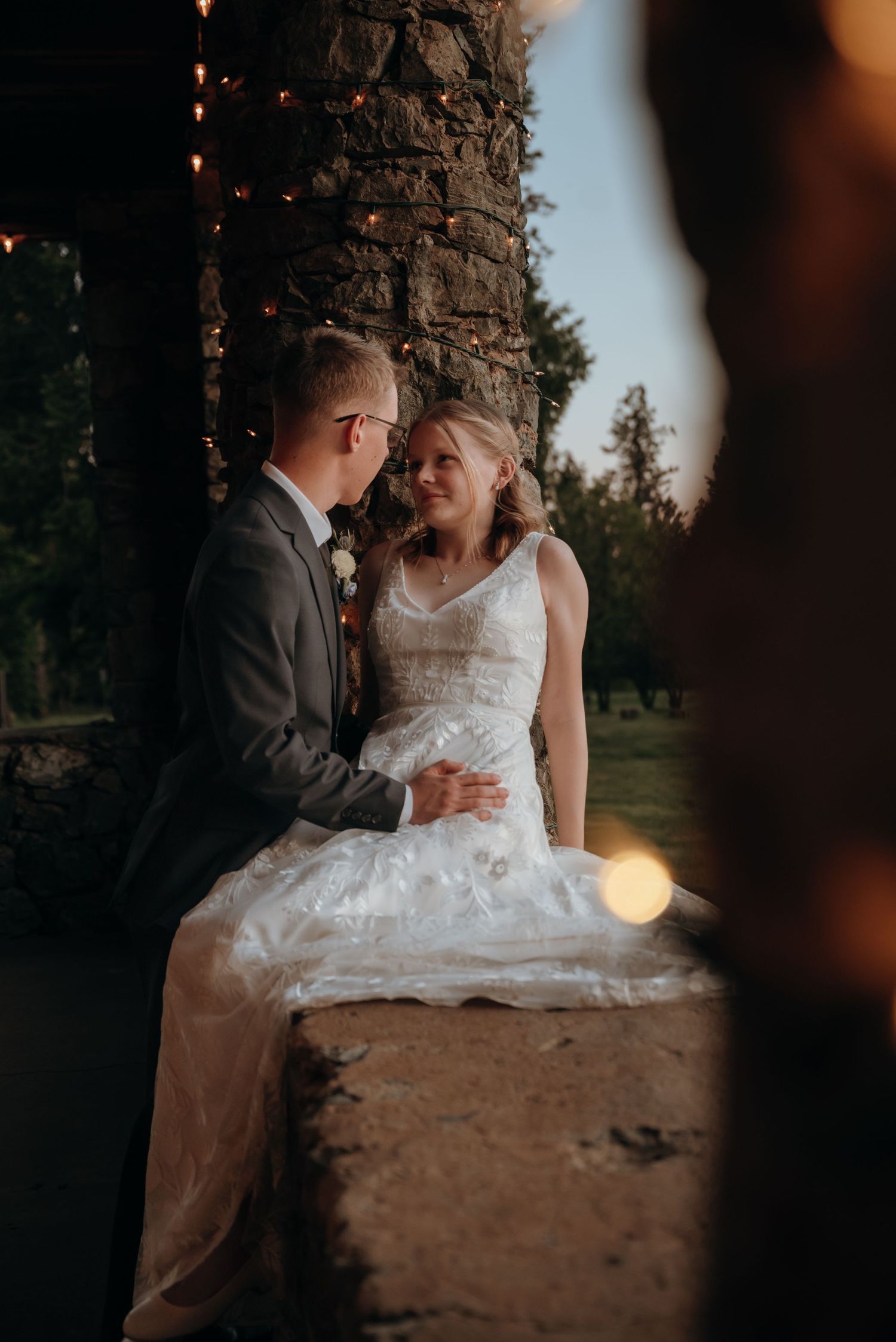 A couple shares a romantic moment on a stone wall at sunset with string lights glowing.