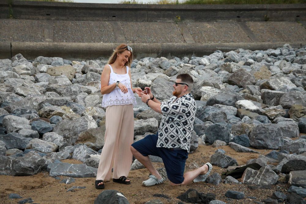 A romantic proposal moment unfolds on a rocky beach shoreline.
