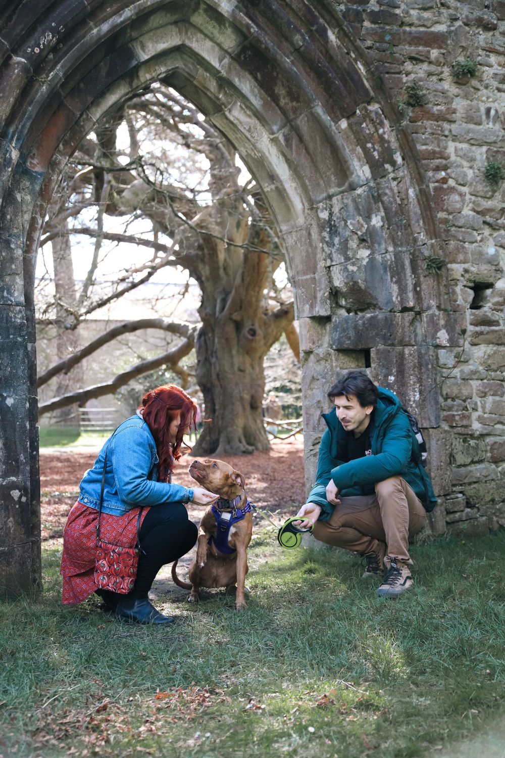 Two people and a dog sit beneath a stone archway of old ruins on a winter day.