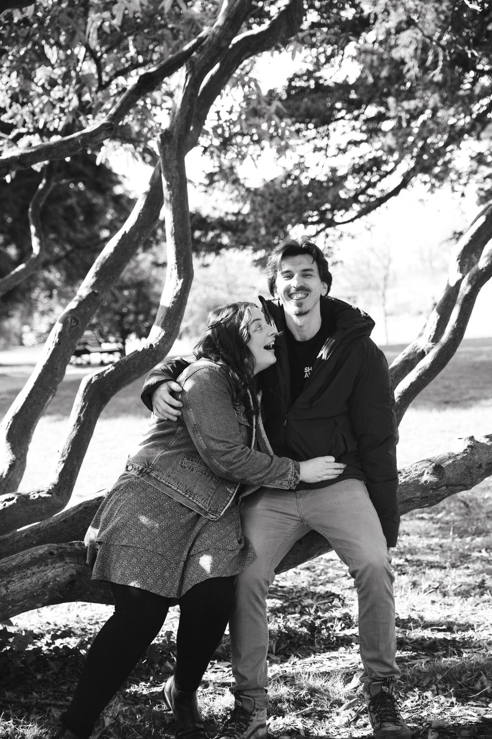 A happy couple laughing together while posing by a twisted tree trunk in a black and white outdoor photo.