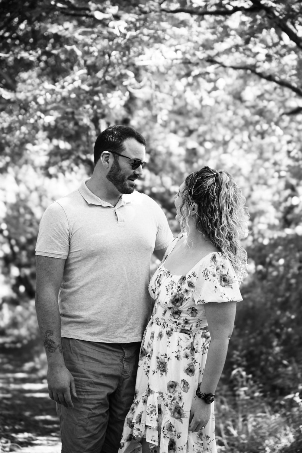 Black and white photo of a couple smiling at each other outdoors, surrounded by lush trees on a sunny day.