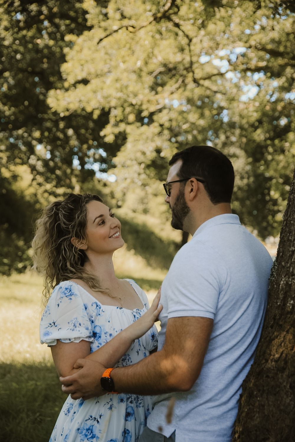 A smiling couple shares a romantic moment by a tree in a sunny park, woman in blue floral dress.