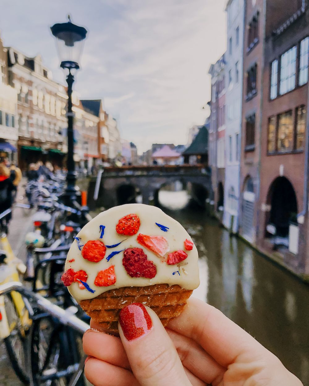 Hand holding white frosted cookie with strawberries along canal in European city.