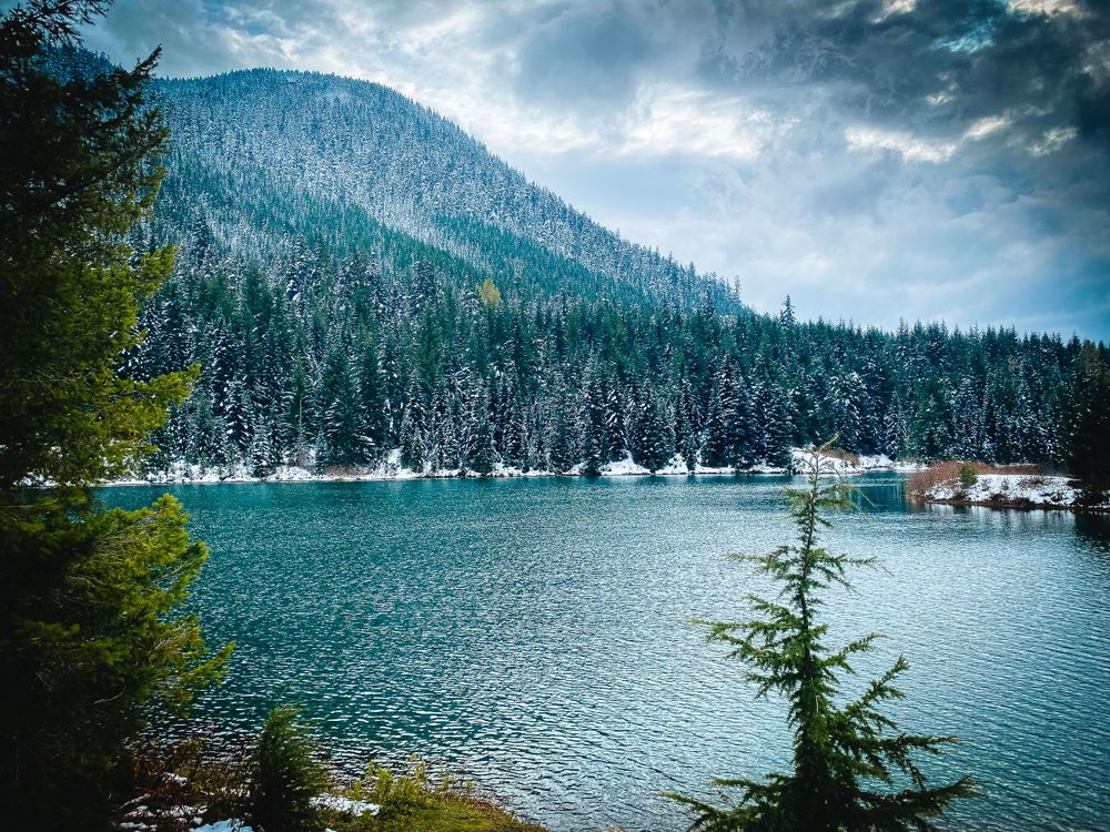 Mountain lake surrounded by evergreen forests and snow-capped peaks under cloudy sky.
