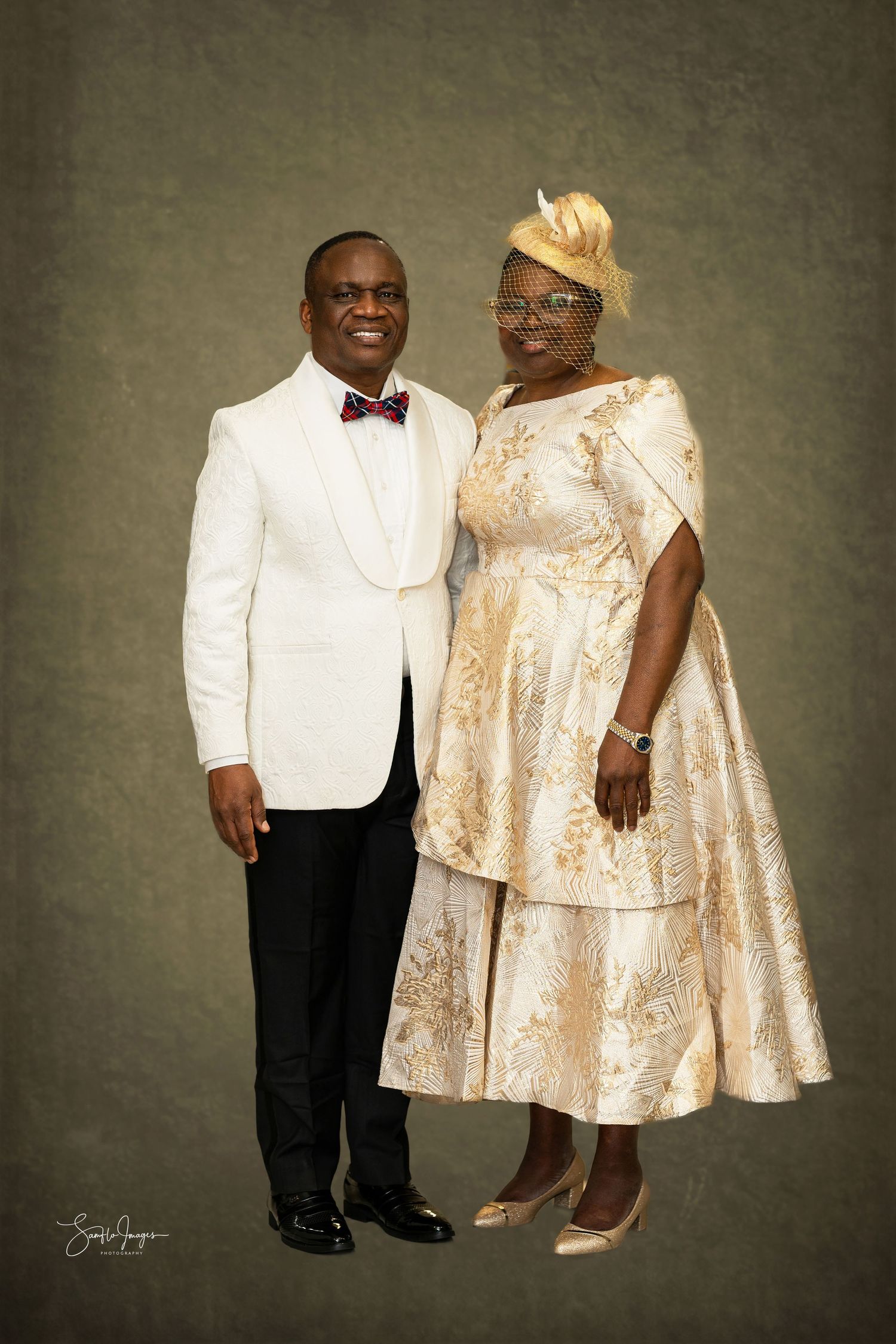 Elegant couple poses in formal attire with white tuxedo jacket and cream-colored floral dress against textured backdrop.
