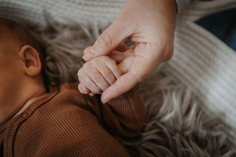 Close-up of tiny baby fingers grasping an adult hand against a soft gray blanket.