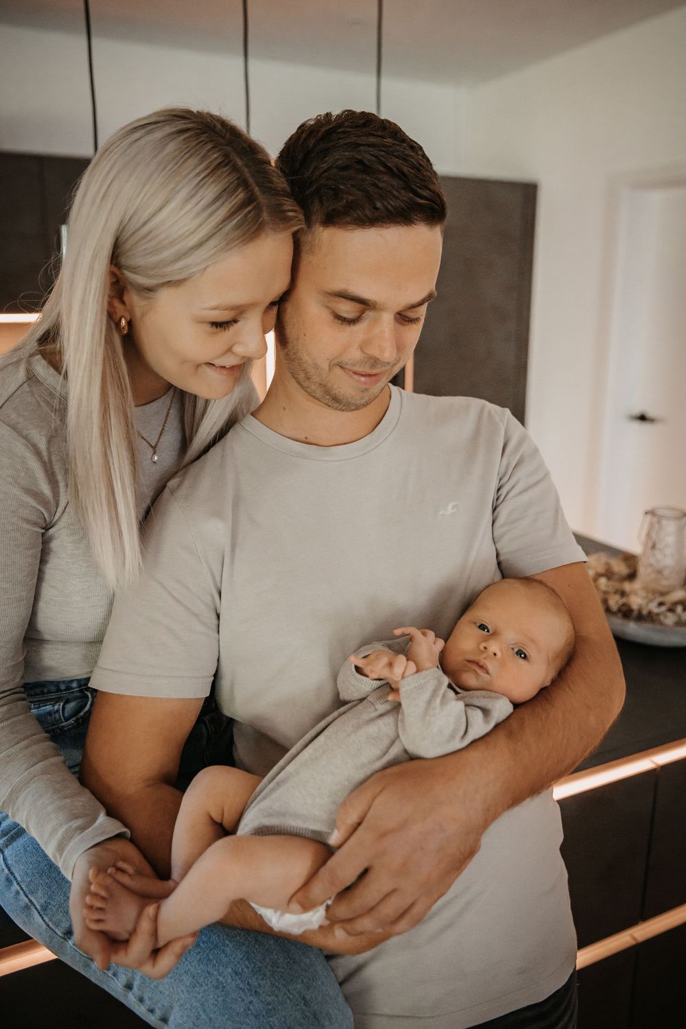 Loving couple in matching gray shirts cuddles with their newborn in natural light.
