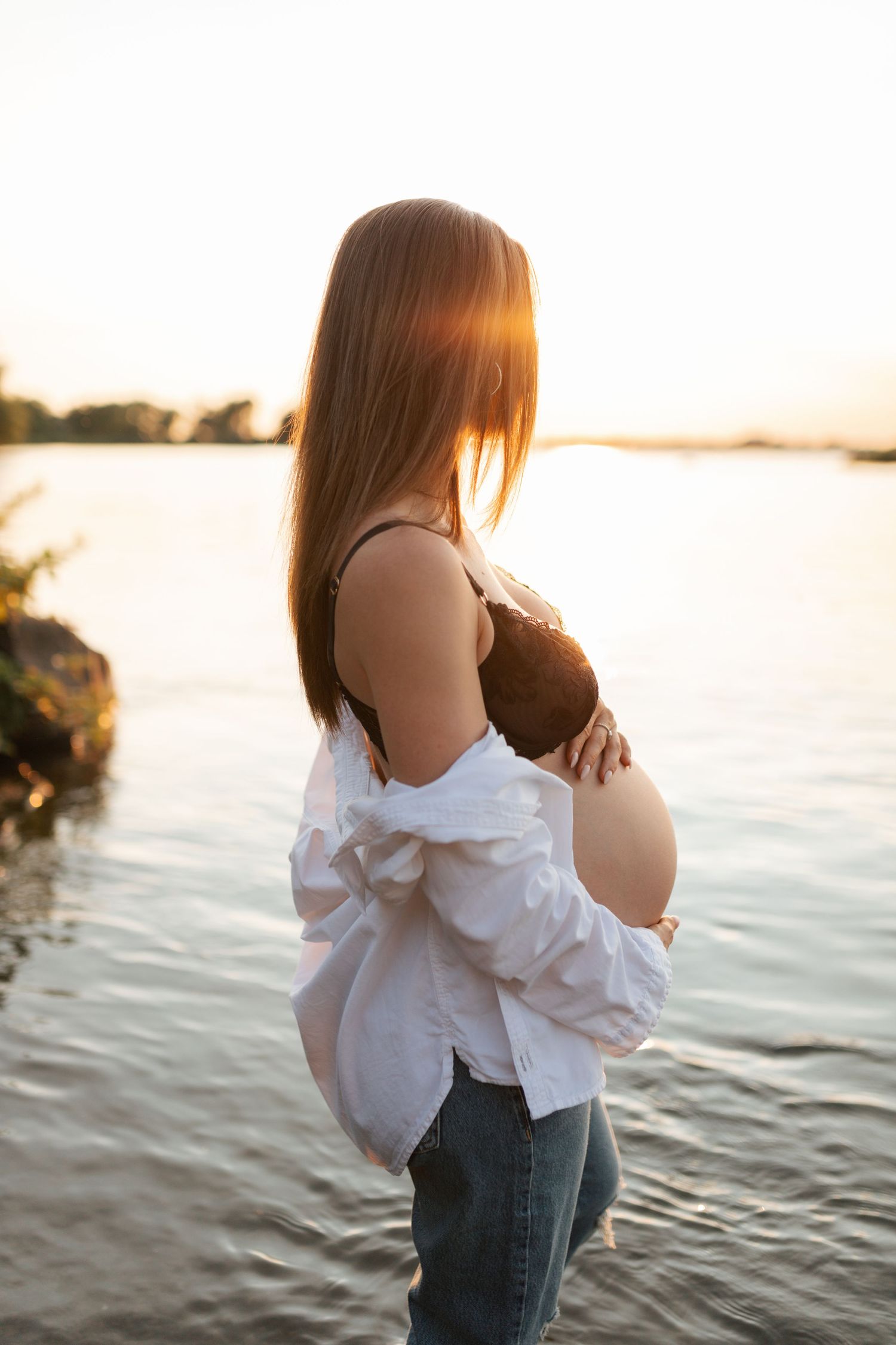 Pregnant woman in white shirt and jeans cradling bare baby bump by sunlit lake at golden hour.
