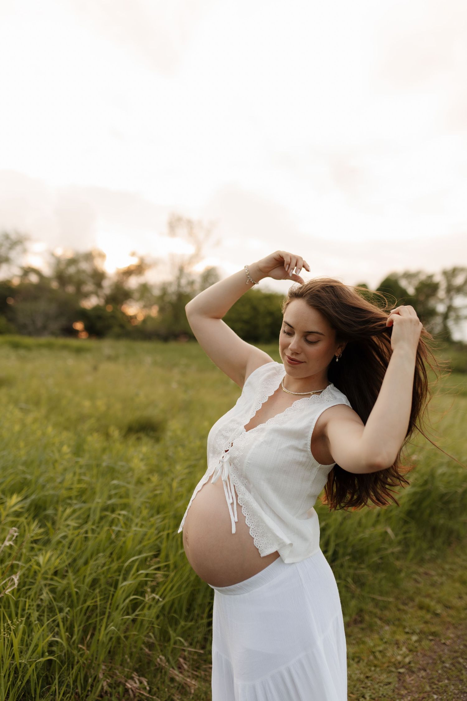 Pregnant woman in white shirt and jeans cradling bare baby bump by sunlit lake at golden hour.