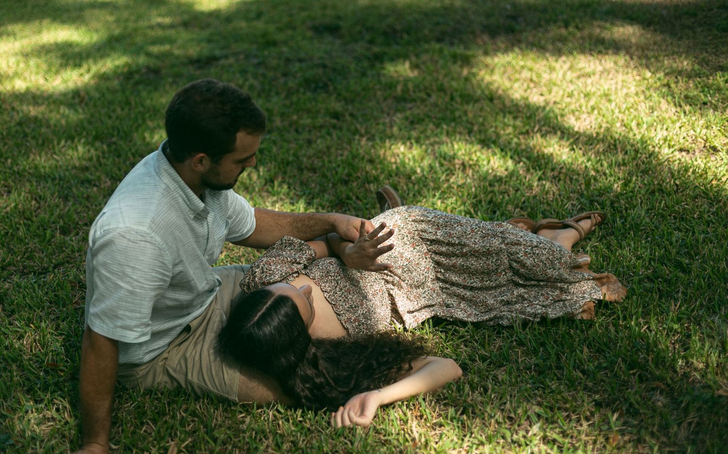 A couple lounges together on a blanket in the grass under dappled sunlight.