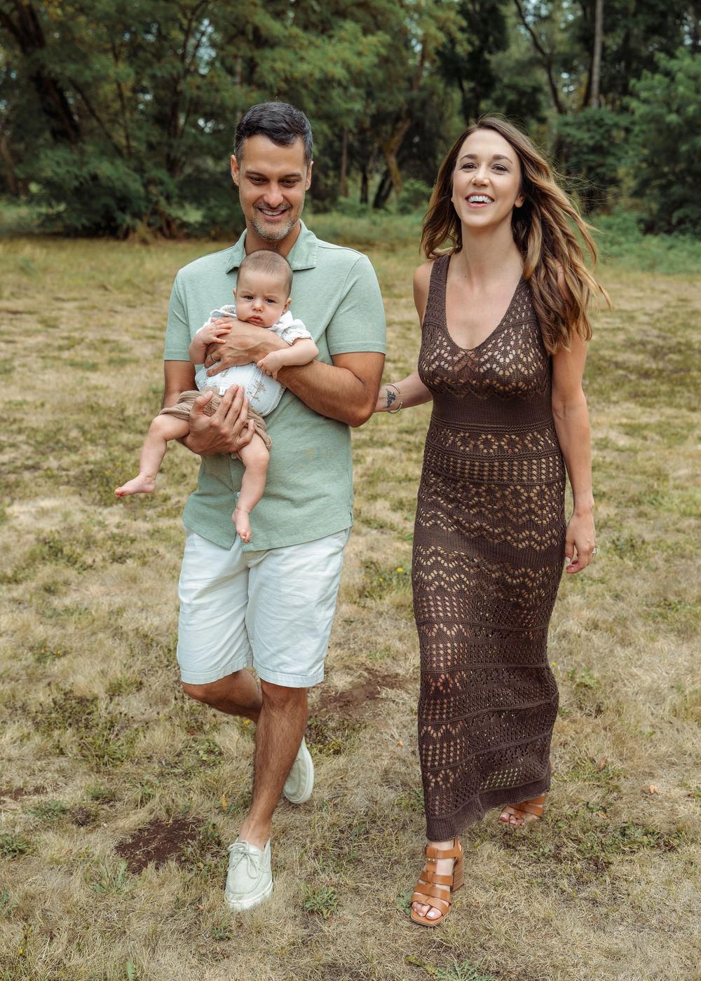 A couple walks together on dry grass while playfully holding and swinging a baby in casual summer outfits.