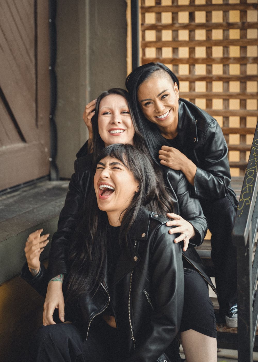 Three friends in black outfits share genuine laughter while sitting together on outdoor steps.