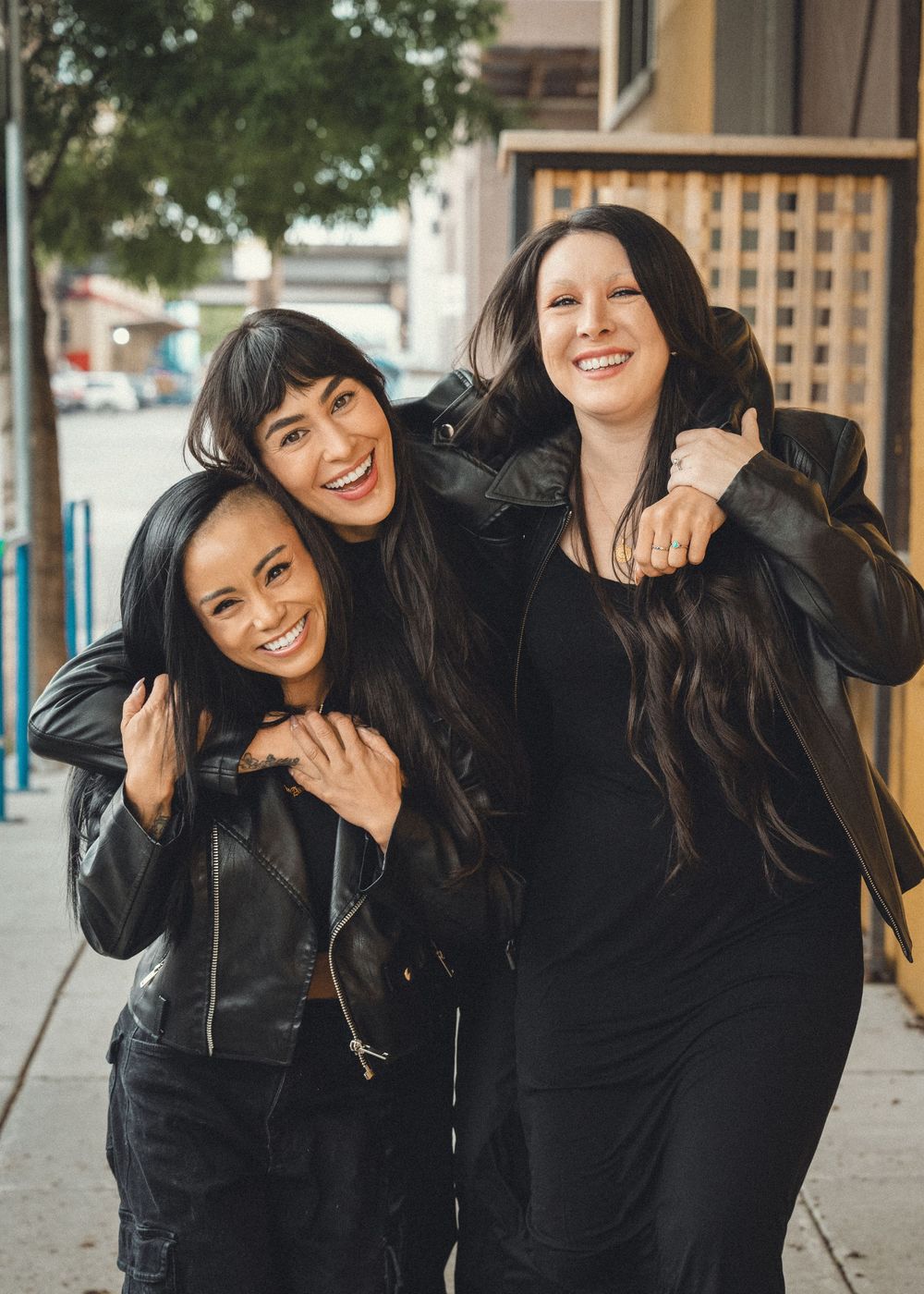 Three friends wearing black leather jackets laugh together while hanging out on a city sidewalk.