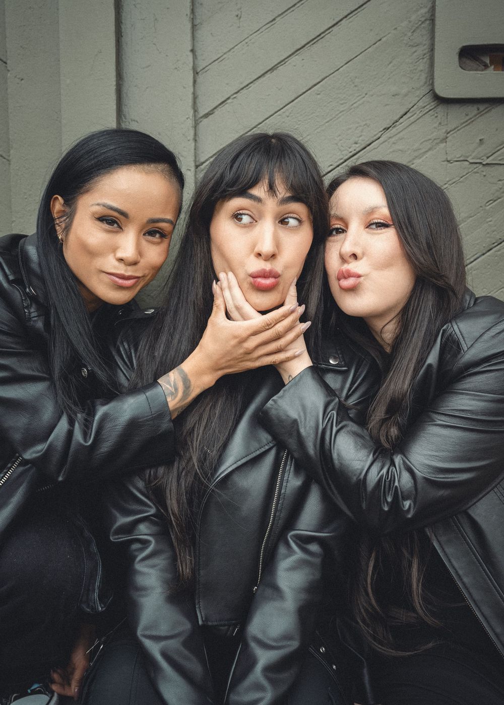 Three friends in black leather jackets strike playful poses against a gray wall.