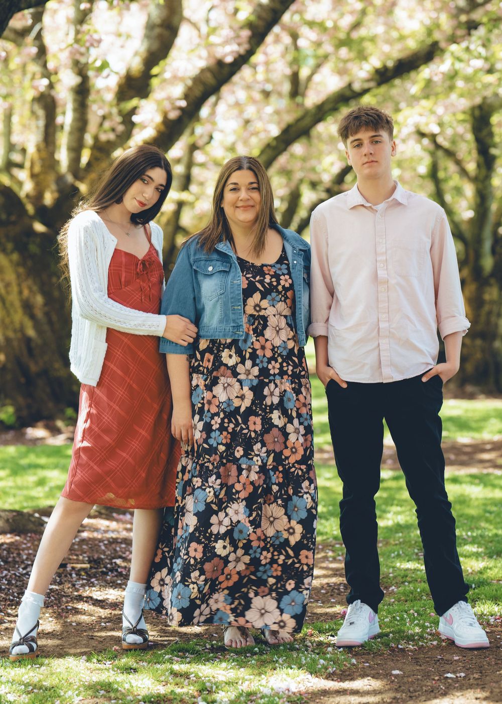 Three people pose together on a dirt path under sunlit trees wearing casual spring outfits in floral and pastel colors.
