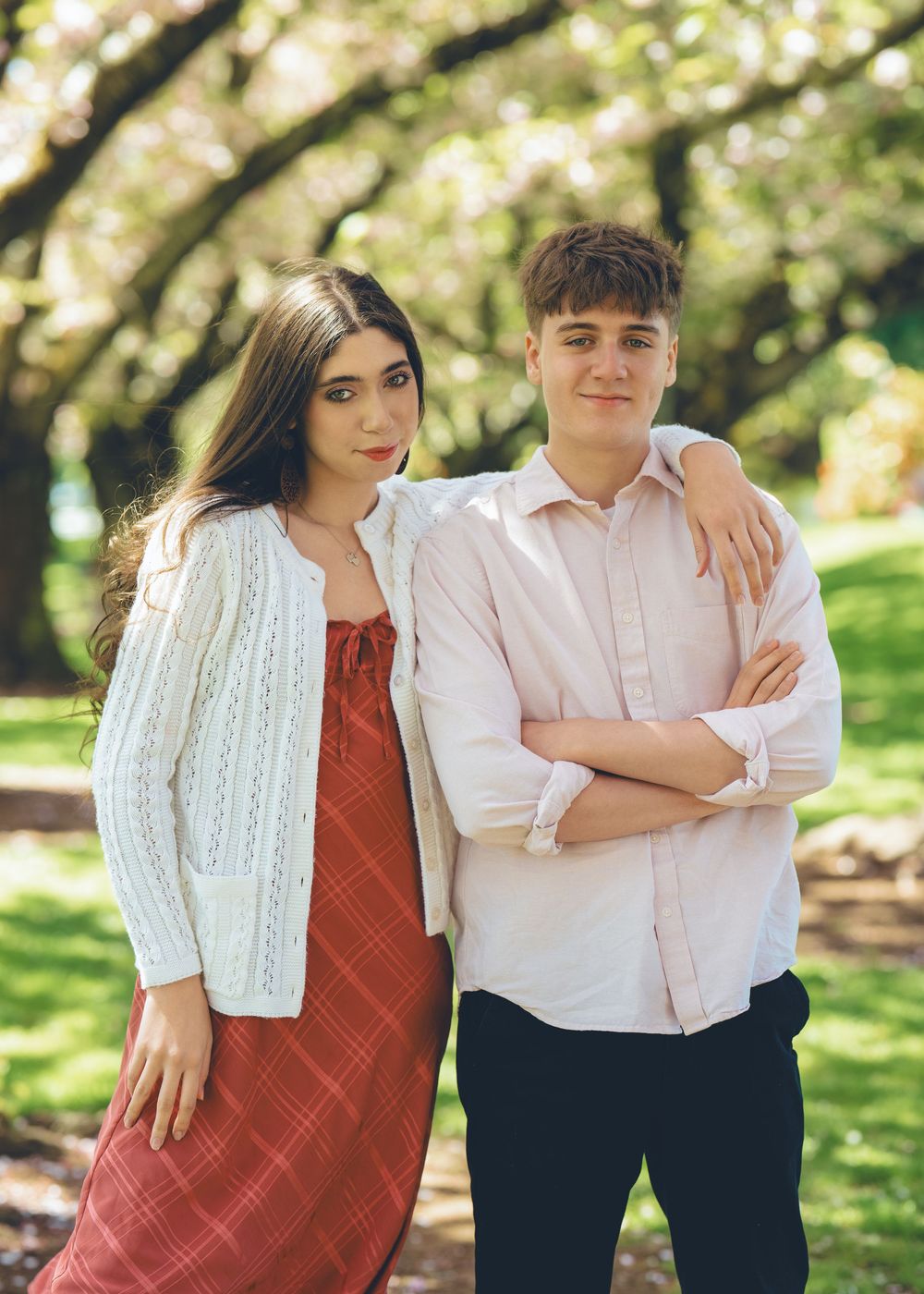 A couple poses together outdoors on a sunny day, with her in a red dress and white sweater and him in a light pink shirt.