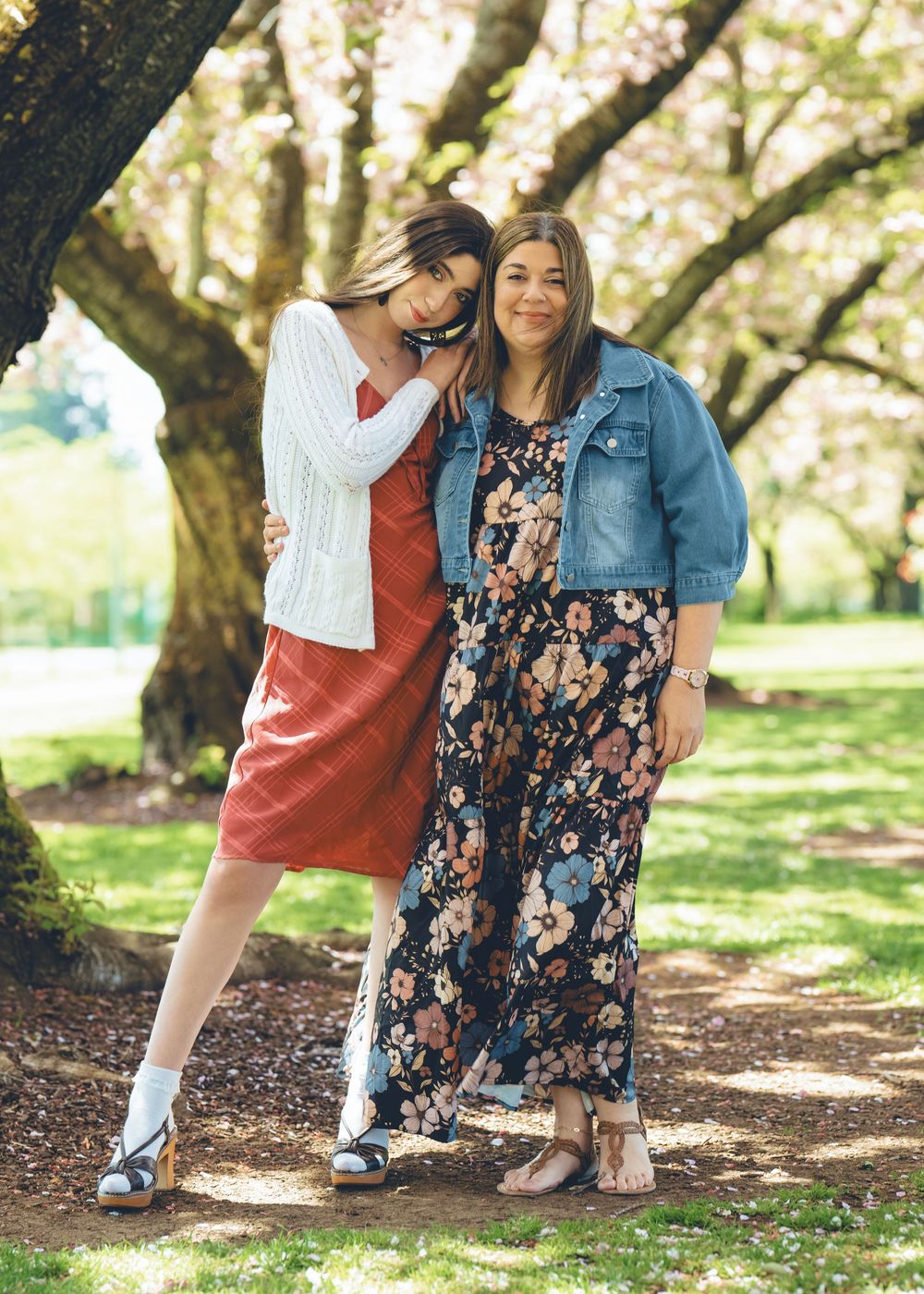 Two friends posing outdoors under cherry blossom trees, wearing casual spring outfits in a park setting.