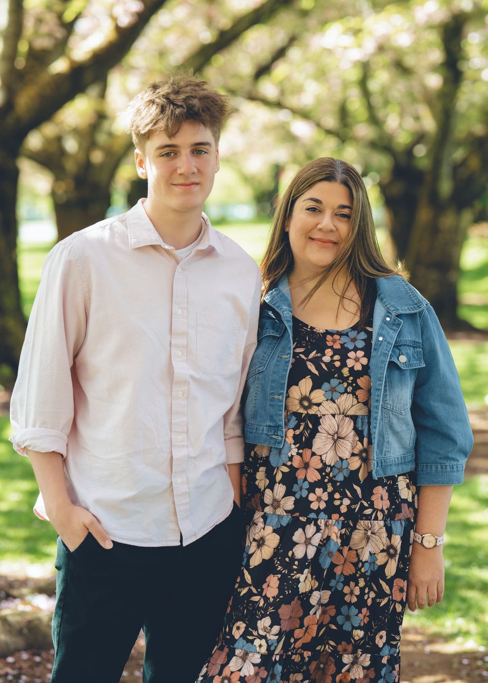 Young couple standing together outdoors in casual spring attire with floral dress and denim jacket against blurred green trees.