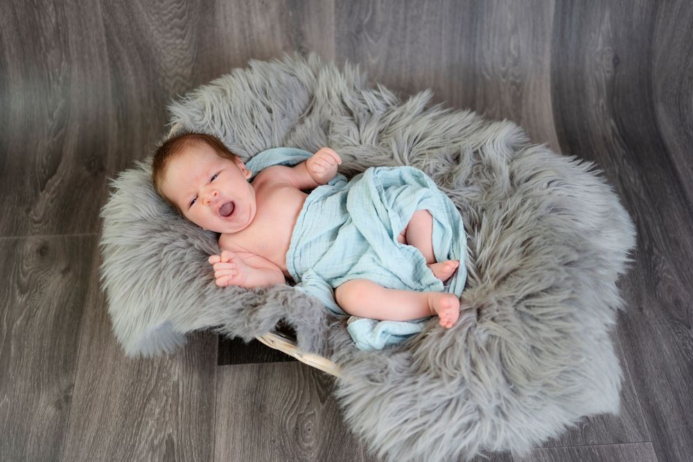Newborn baby yawning while lying on a fluffy gray fur rug, wrapped in a soft blue blanket inside a wooden box.