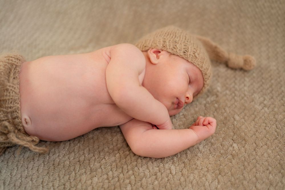 Sleeping newborn baby wearing a knit beige hat and shorts outfit, lying on a soft tan carpet.