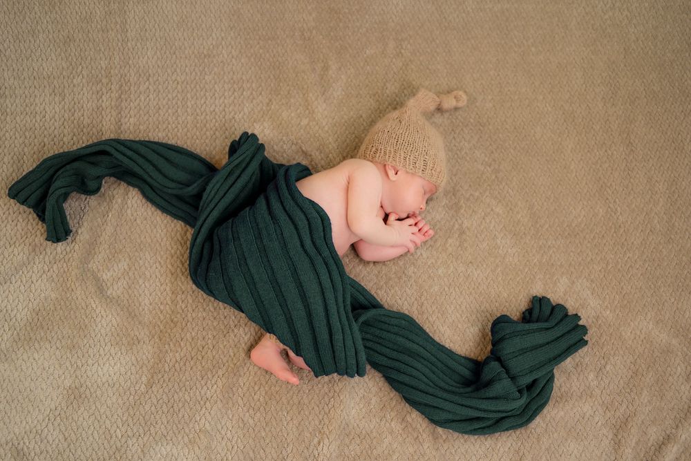 Newborn baby sleeping peacefully wrapped in a dark green knit blanket wearing a beige knot hat on a tan background.