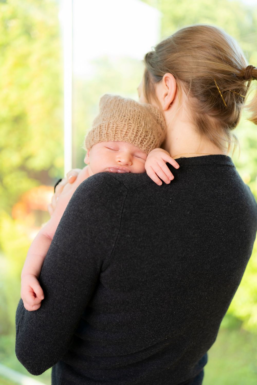 Mother holding sleeping newborn baby in knit hat against her shoulder near a bright window.