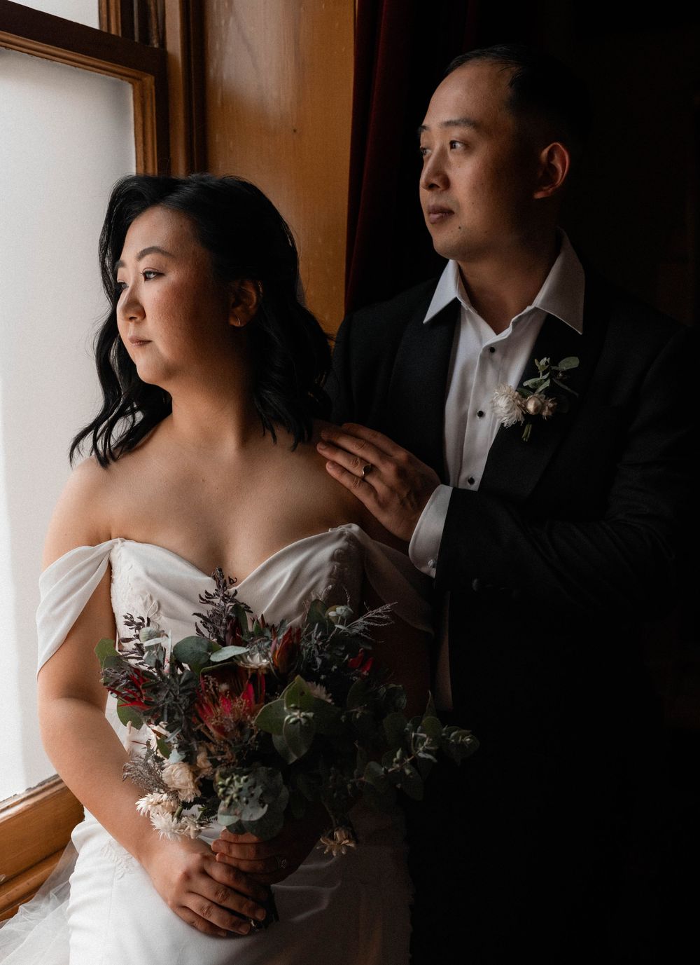 Bride in white strapless dress holds moody burgundy bouquet while standing by window with groom in black suit.