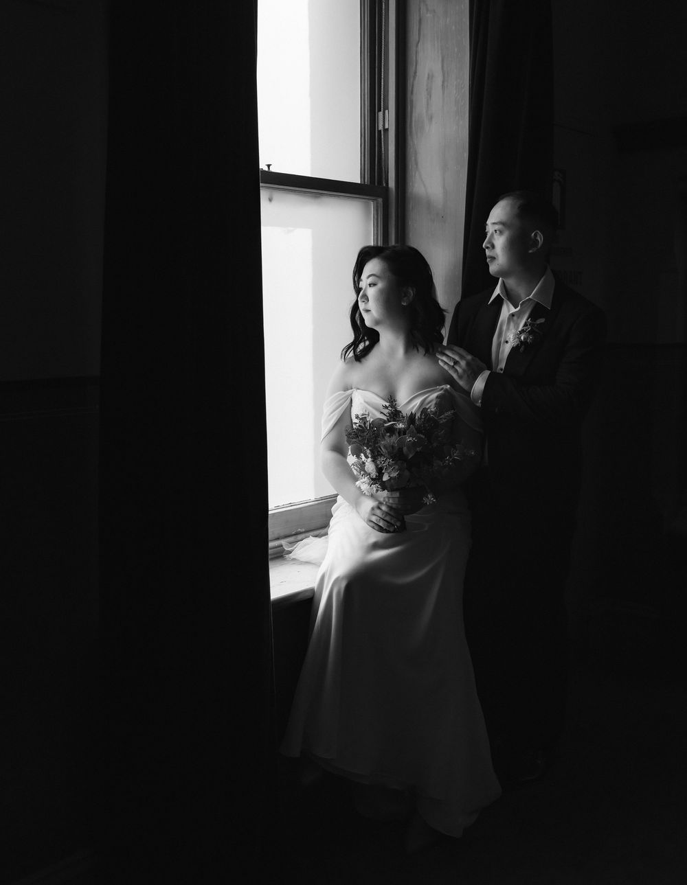 A dramatic black and white wedding portrait shows a couple in elegant attire standing by a window in moody lighting.