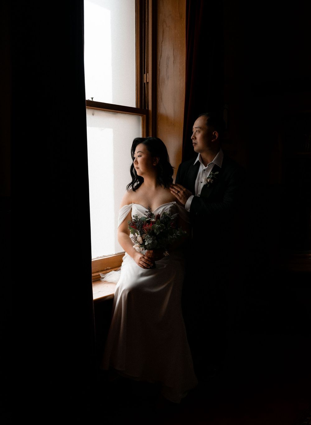 A bride in a white wedding dress stands near a window, creating a dramatic silhouette against natural light.