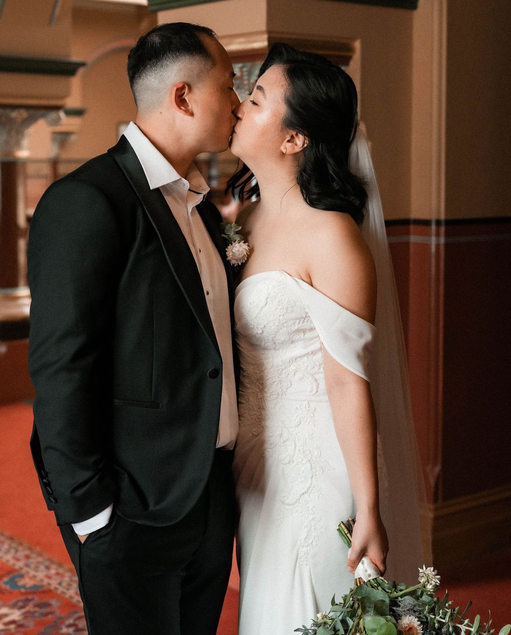A romantic wedding couple shares intimate moments in an elegant indoor venue with warm lighting and floral bouquet details.