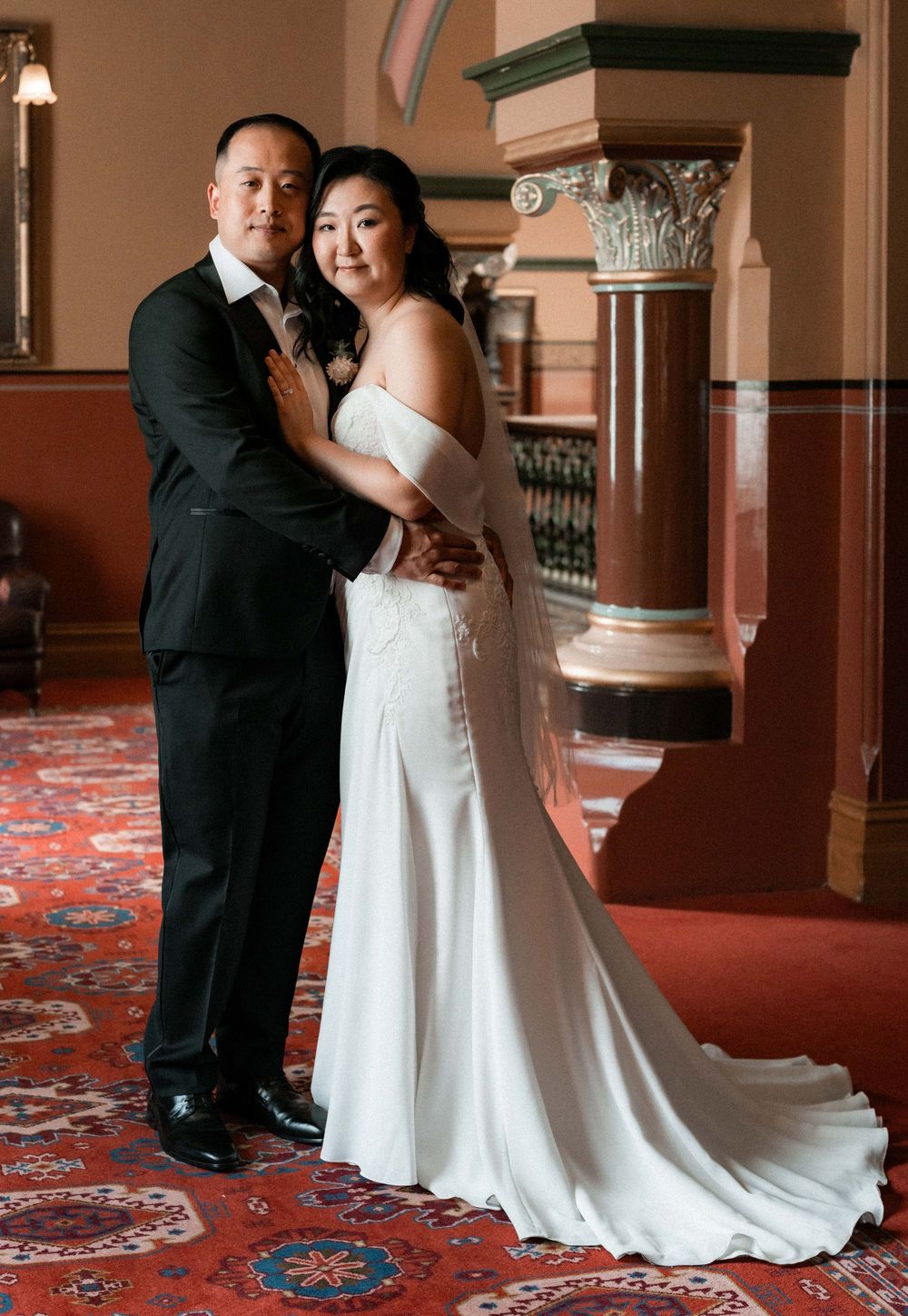 A couple in wedding attire dances together on a patterned red carpet in an elegant venue with ornate columns.