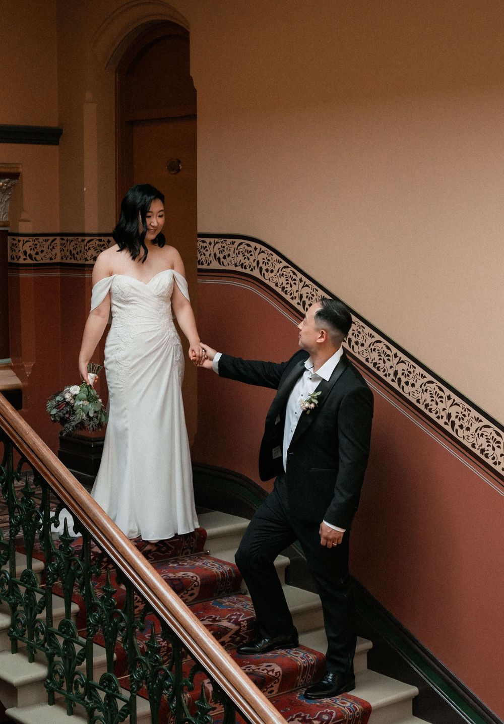 A couple shares an intimate moment on ornate carpeted stairs with decorative railings in a dimly lit hallway.
