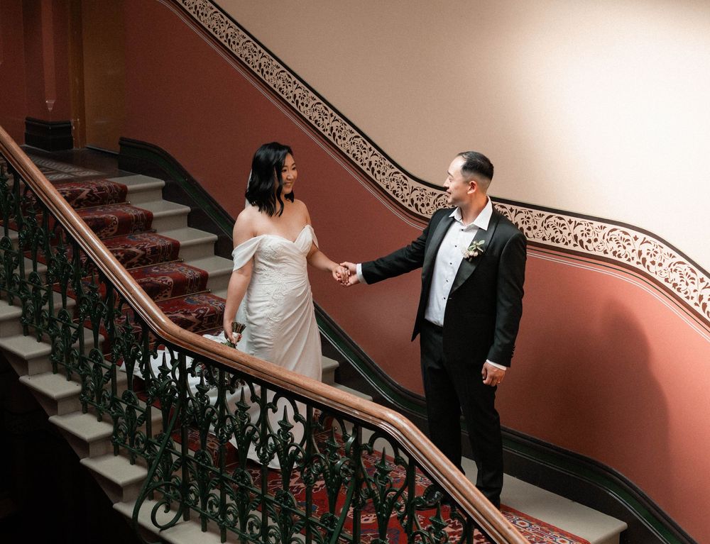 A couple ascends an elegant staircase with ornate iron railings and coral-colored walls in a romantic setting.